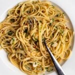 Spaghetti on a white plate in a aglio e olio sauce. Sliced garlic and parsley mixed through a a fork resting in.