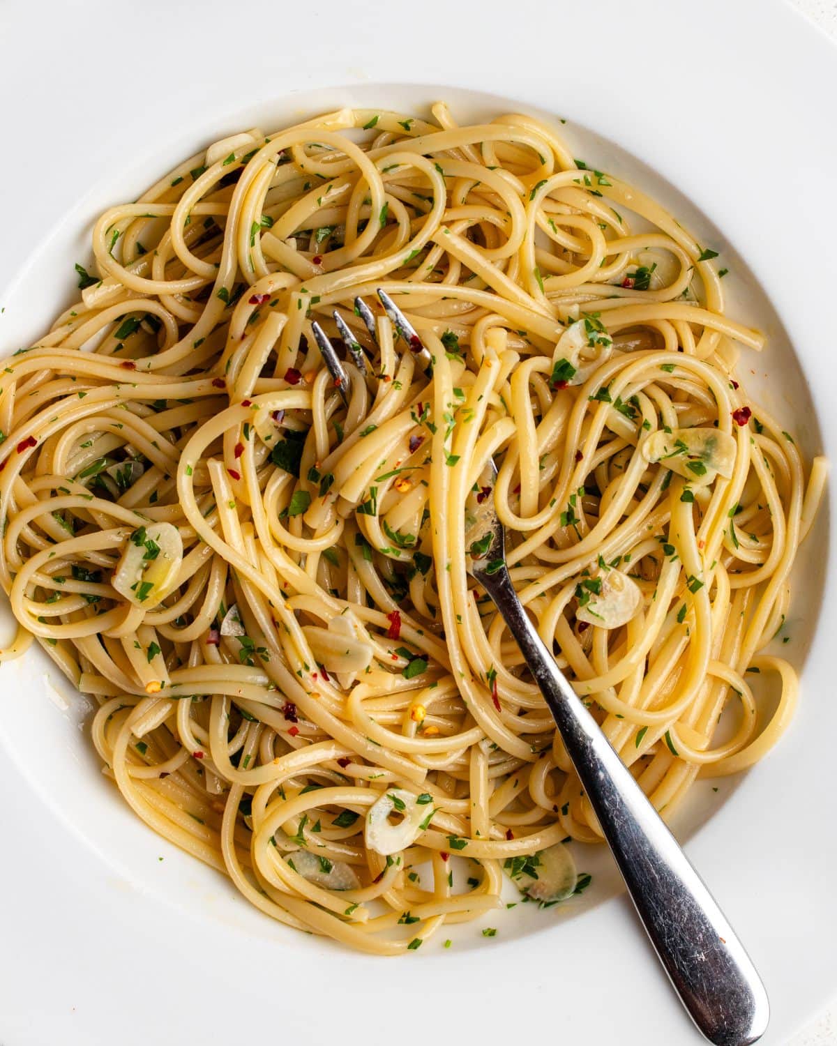 Spaghetti on a white plate in a aglio e olio sauce. Sliced garlic and parsley mixed through a a fork resting in.