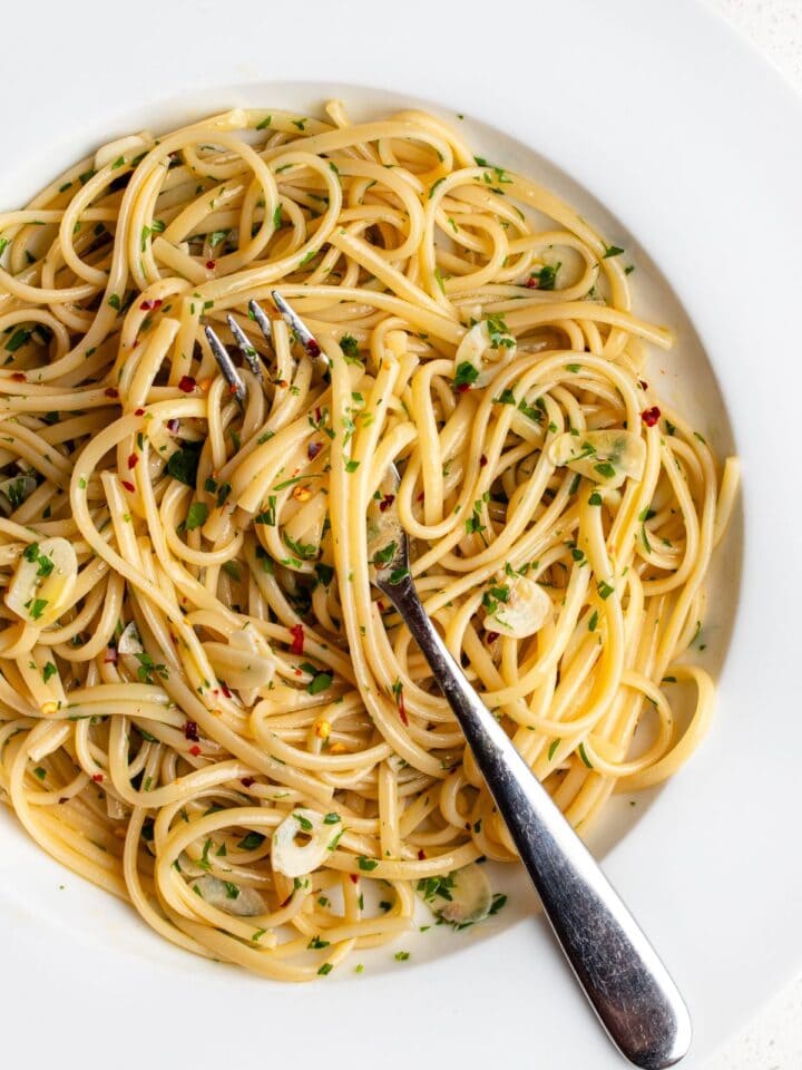 White plate with spaghetti in an aglio e olio sauce. Fork resting in and chopped parsley on top.