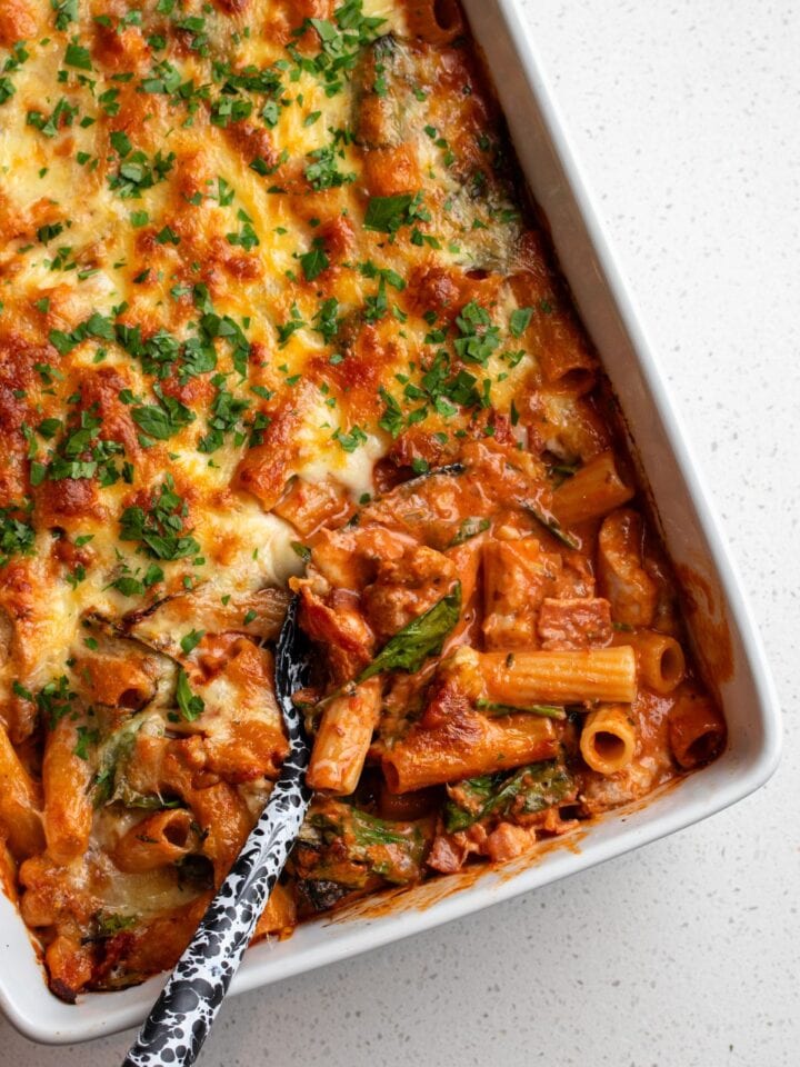 A white porcelain dish with a chicken pasta bake. A spoon is digging in to show the creamy tomato sauce mixed in to pasta. Golden cheese on top.
