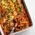 A white porcelain dish with a chicken pasta bake. A spoon is digging in to show the creamy tomato sauce mixed in to pasta. Golden cheese on top.
