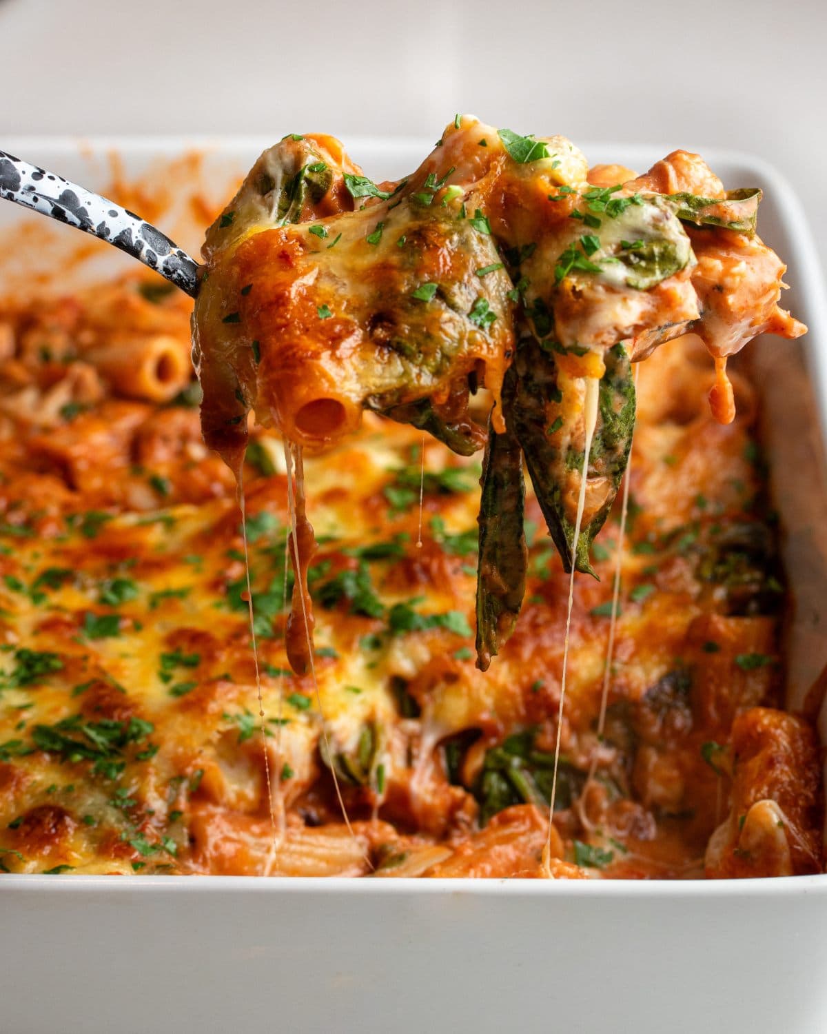 A portion of the pasta bake being lifted out of the dish with a spoon. Stretchy mozzarella pulling away.