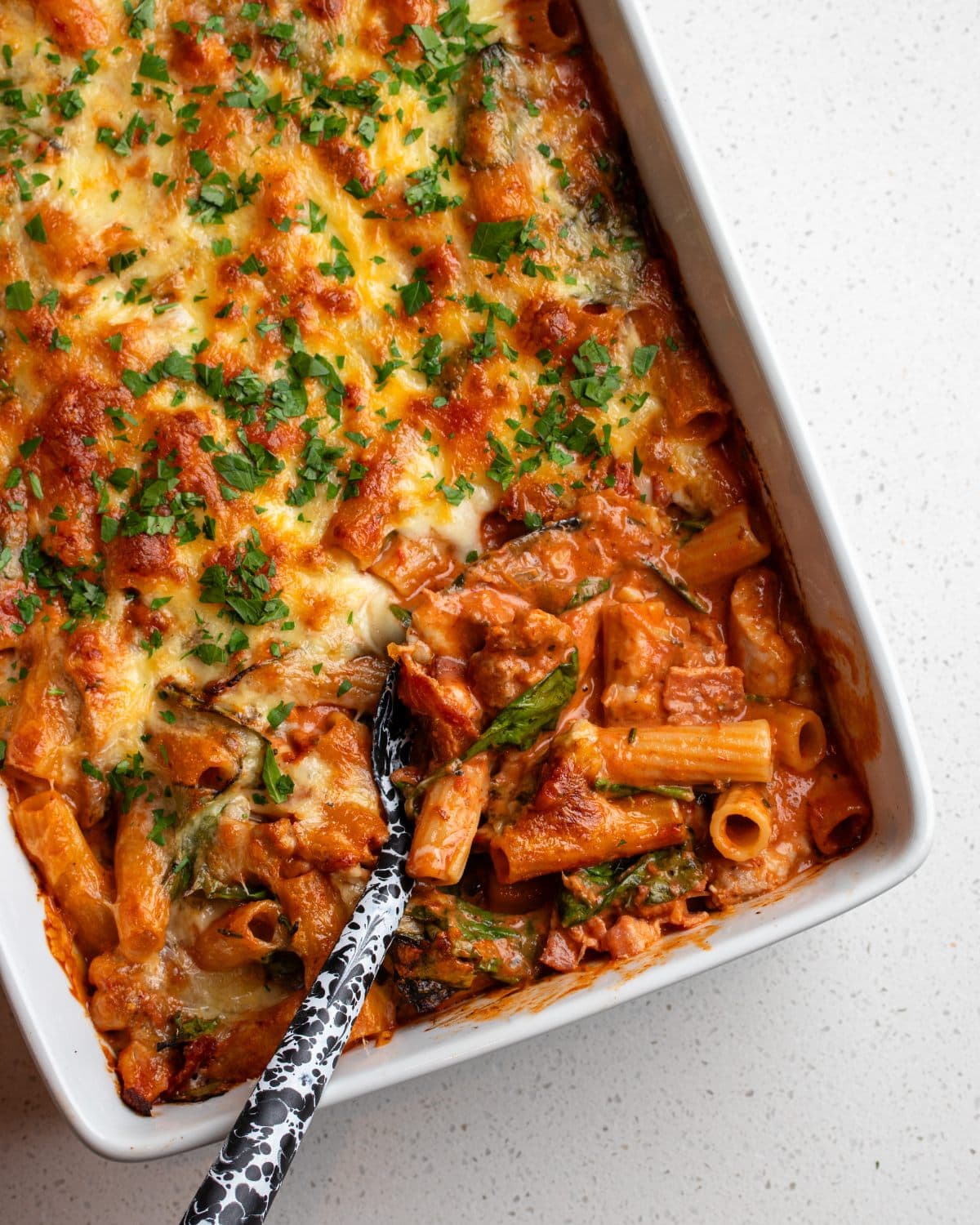 A white porcelain dish with a chicken pasta bake. A spoon is digging in to show the creamy tomato sauce mixed in to pasta. Golden cheese on top.