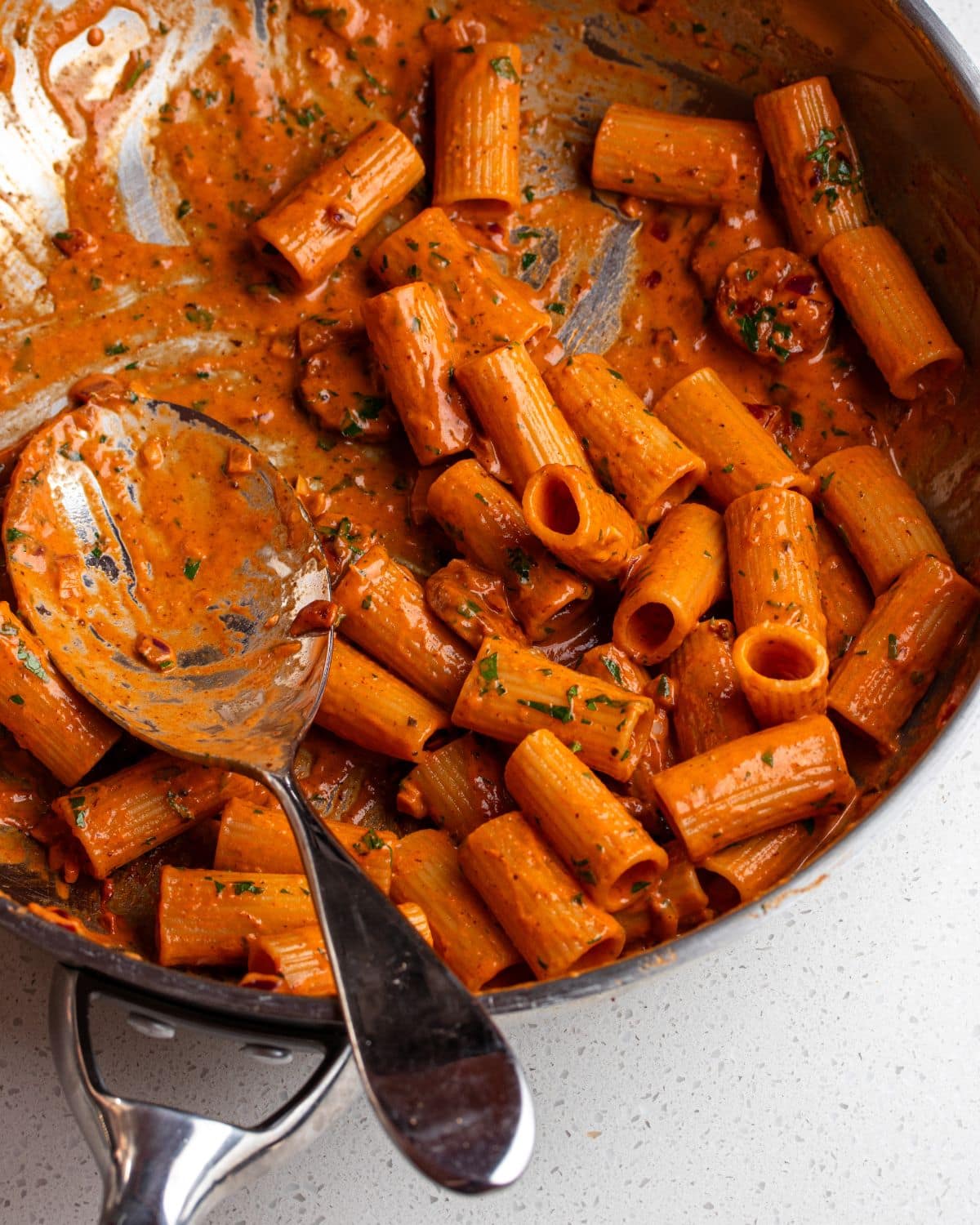Rigatoni in the creamy chorizo sauce in a stainless steel saucepan with a serving spoon resting in.