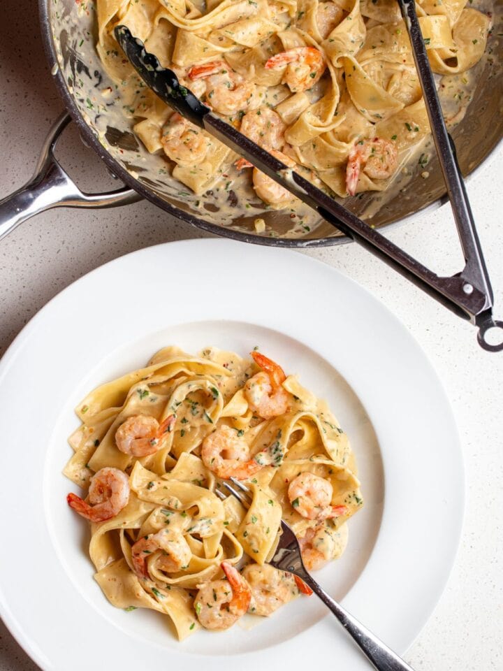 Bench top with a white plate with creamy shrimp pasta and the fry pan above with remaining pasta and tongs resting in.