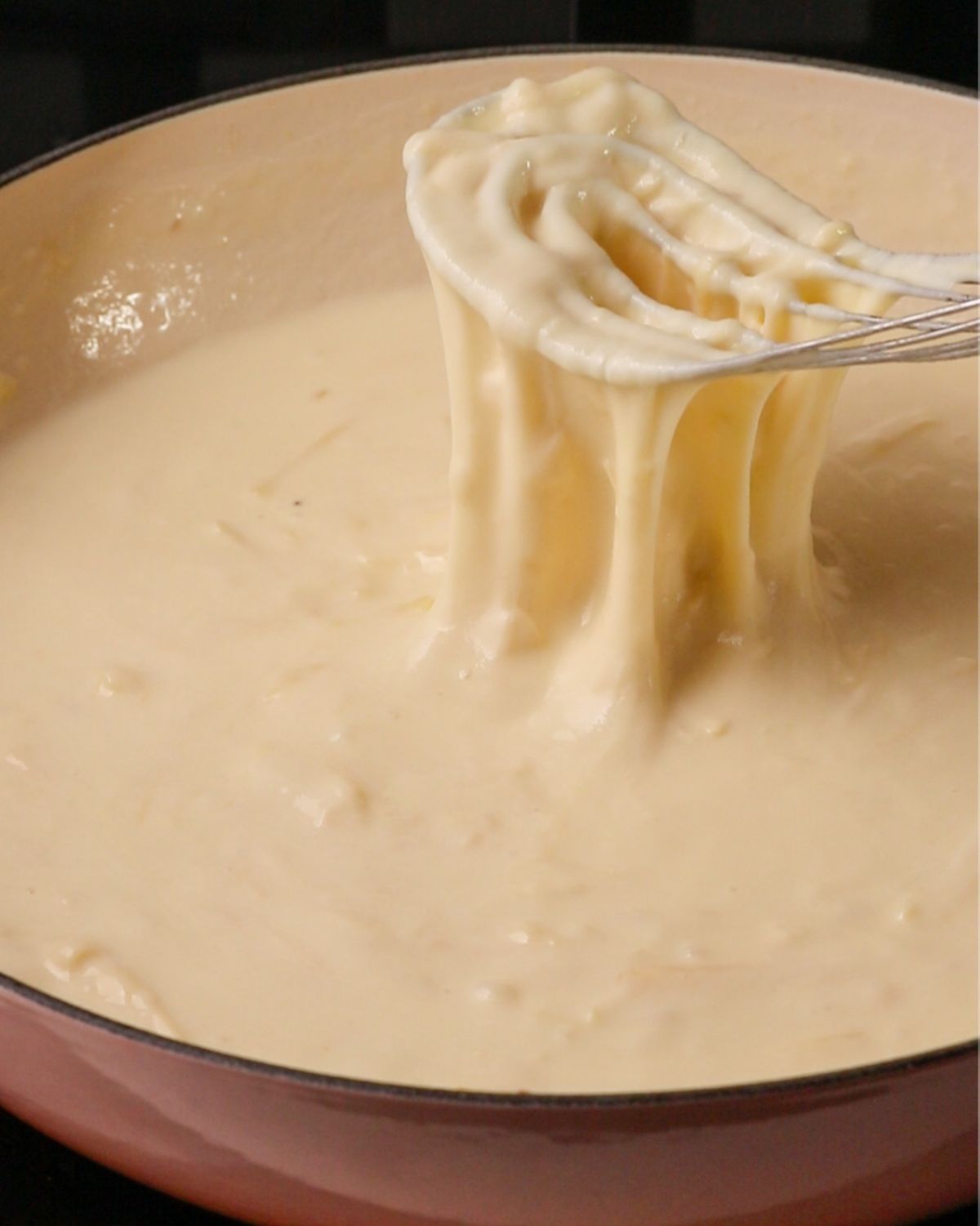 A whisk mixing a cheese sauce with cheese pulling off the whisk.