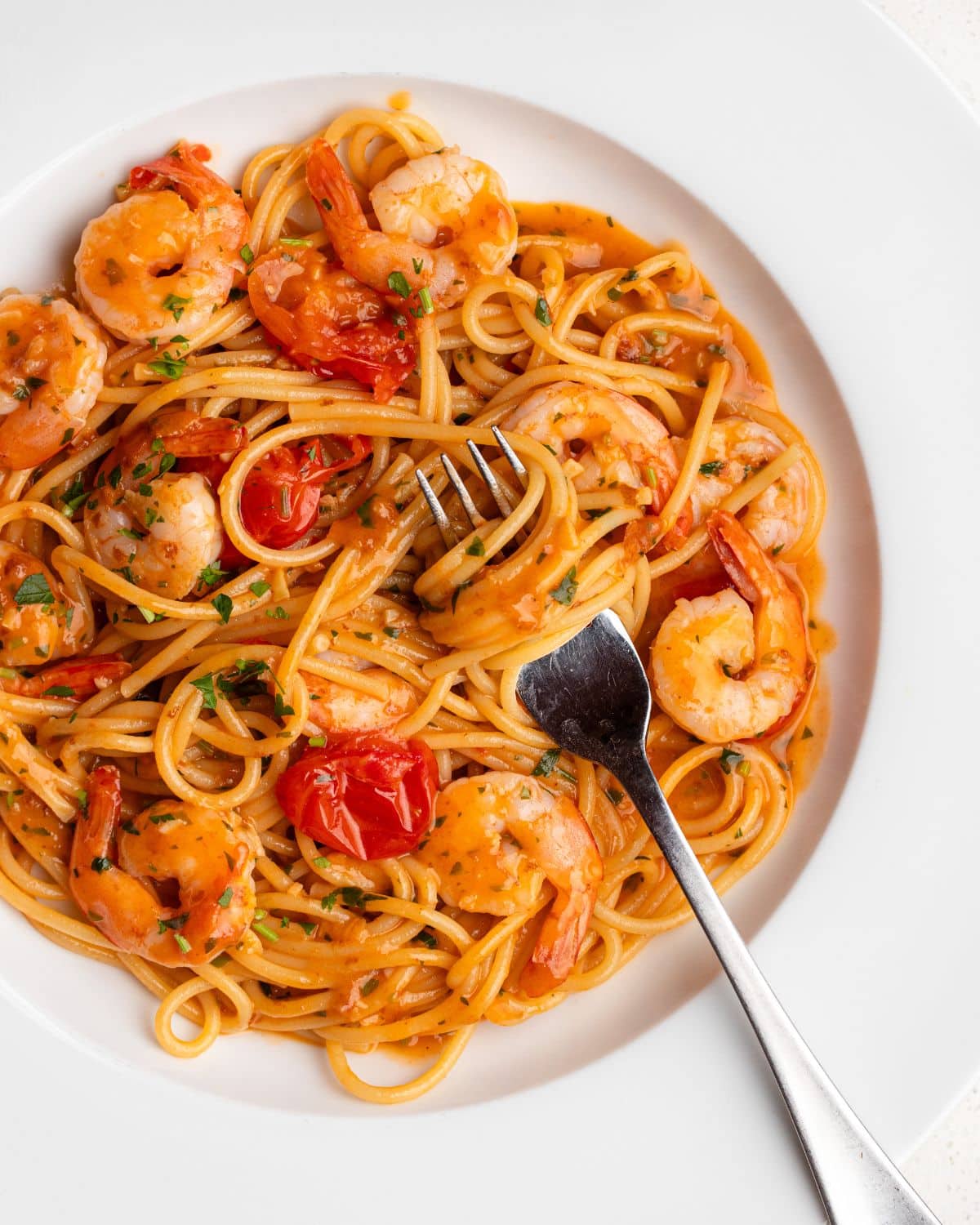 White plate with shrimp pasta and a fork twirling noodles. Cherry tomatoes and parsley on top.