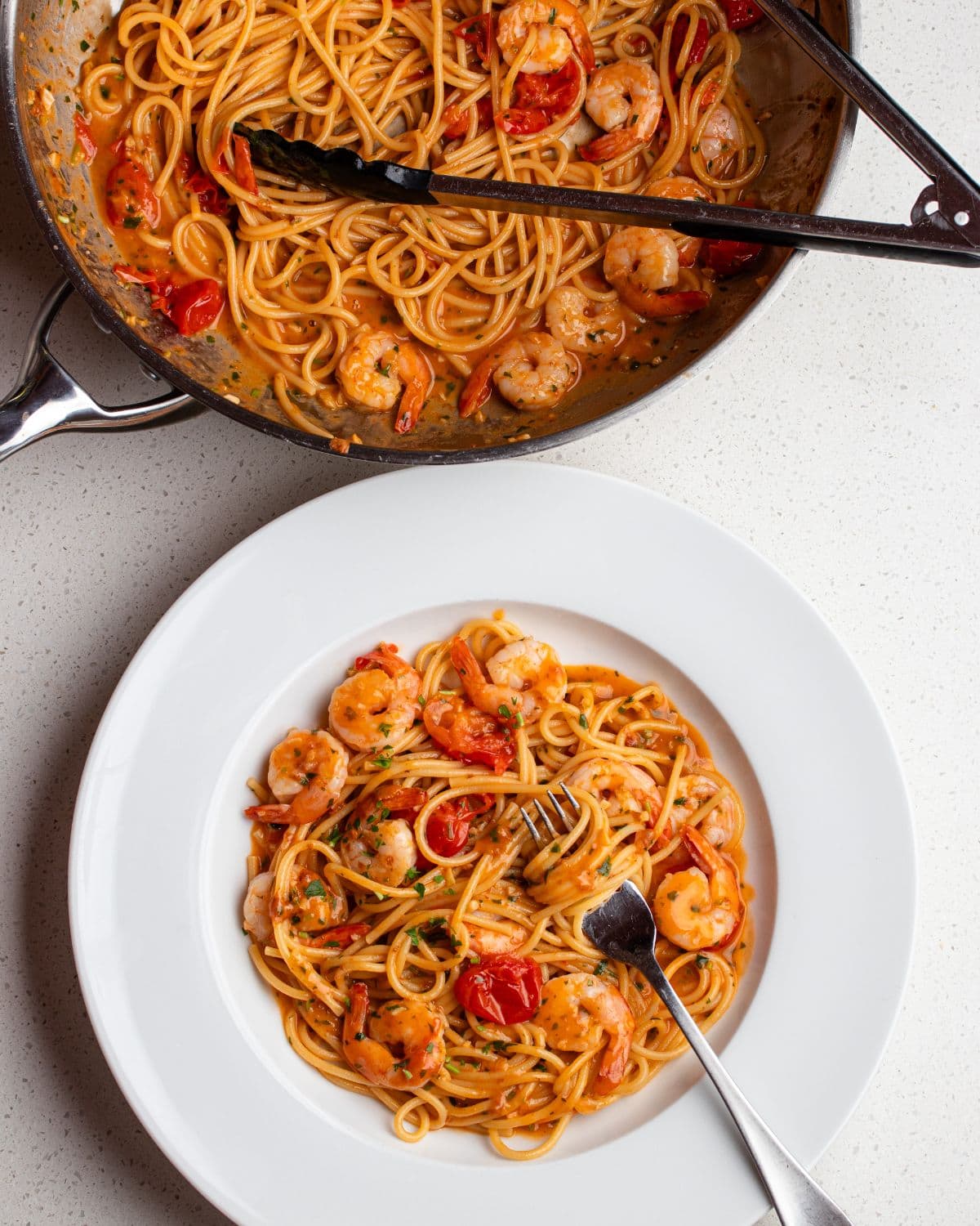White bench top with plate holding shrimp pasta and a fry pan above with remaining pasta.