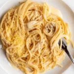 A white plate with spaghetti in a creamy white pasta sauce. A fork resting with noodles wrapped around it.