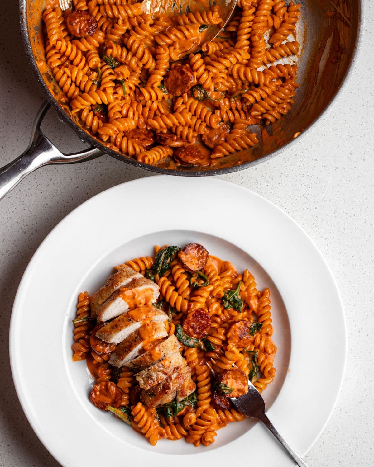 Bench top with finished chicken chorizo pasta with a fork resting in. Above the stainless steel fry pan with remaining pasta.