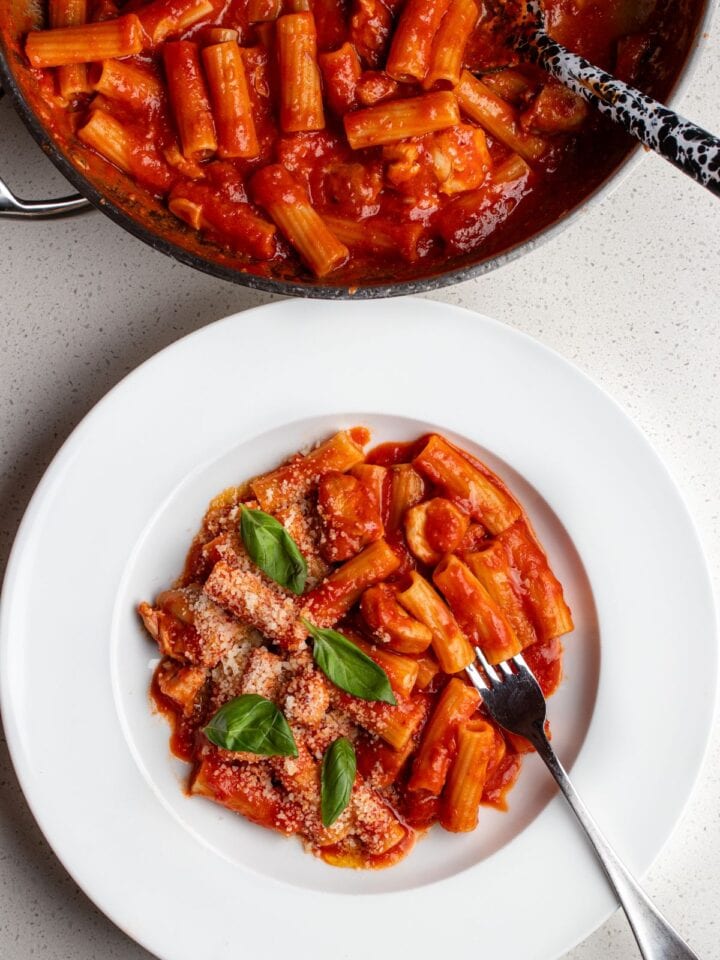 Bench top with a plate of spicy chicken pasta and the fry pan above with remaining pasta and serving spoon.