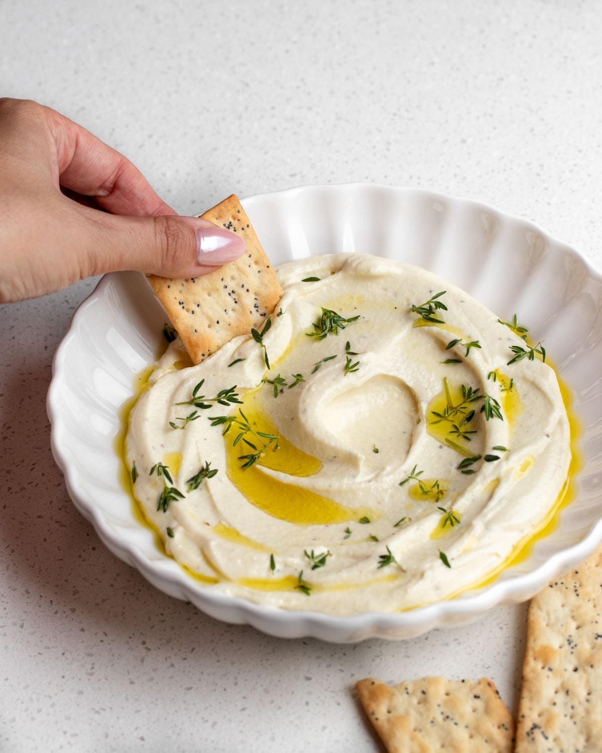 A cracker being dipped into a bowl of whipped ricotta. Olive oil and thyme leaves on top.