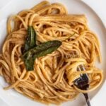 Linguine in a butter parmesan sauce in a white bowl with a fork resting in. Crisp sage leaves on top.
