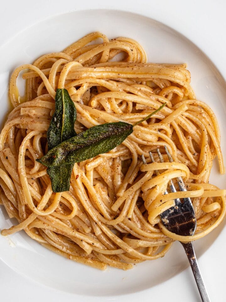 Linguine in a butter parmesan sauce in a white bowl with a fork resting in. Crisp sage leaves on top.