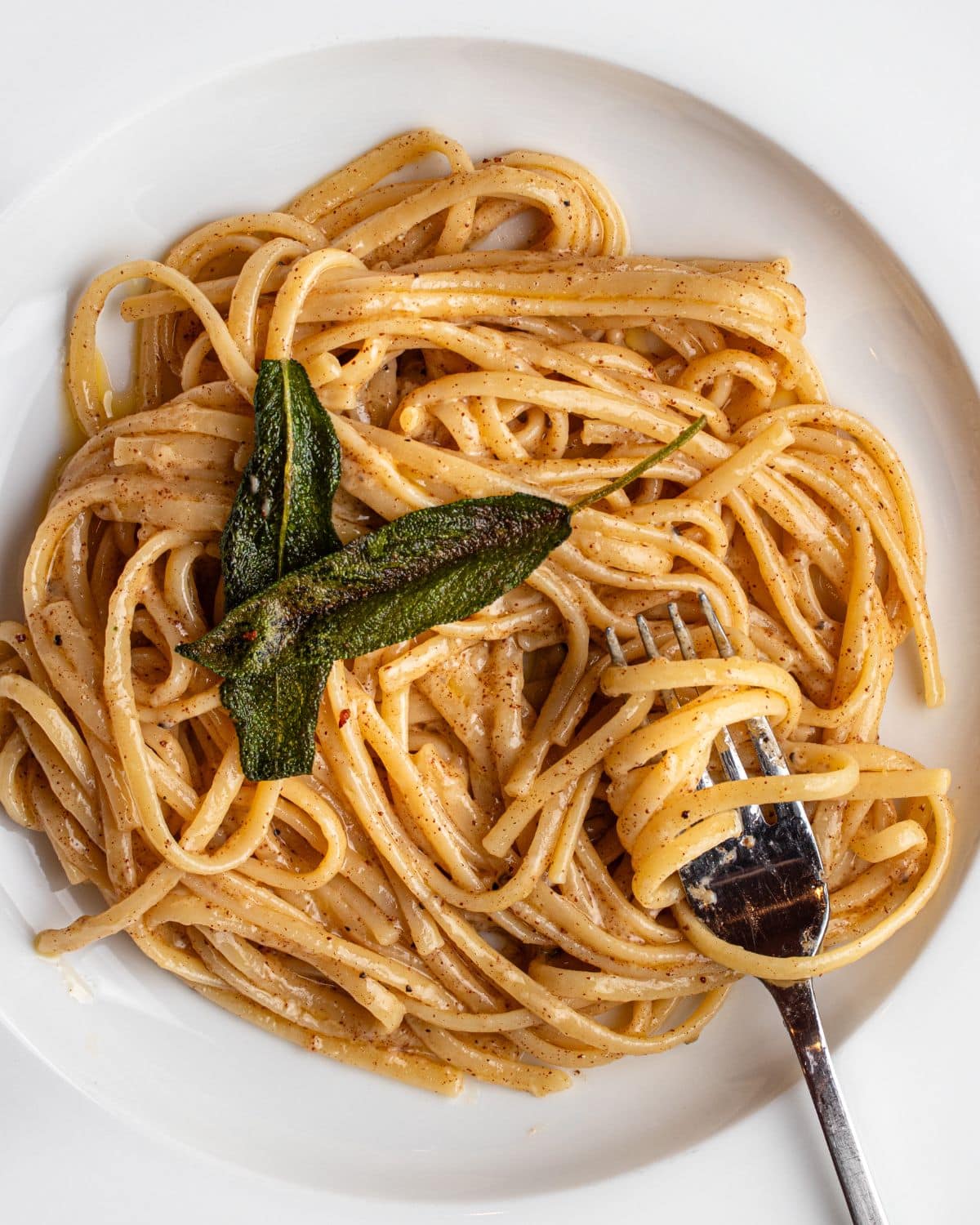 Linguine in a butter parmesan sauce in a white bowl with a fork resting in. Crisp sage leaves on top.