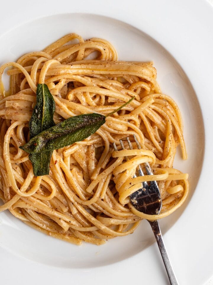 Linguine in a butter parmesan sauce in a white bowl with a fork resting in. Crisp sage leaves on top.