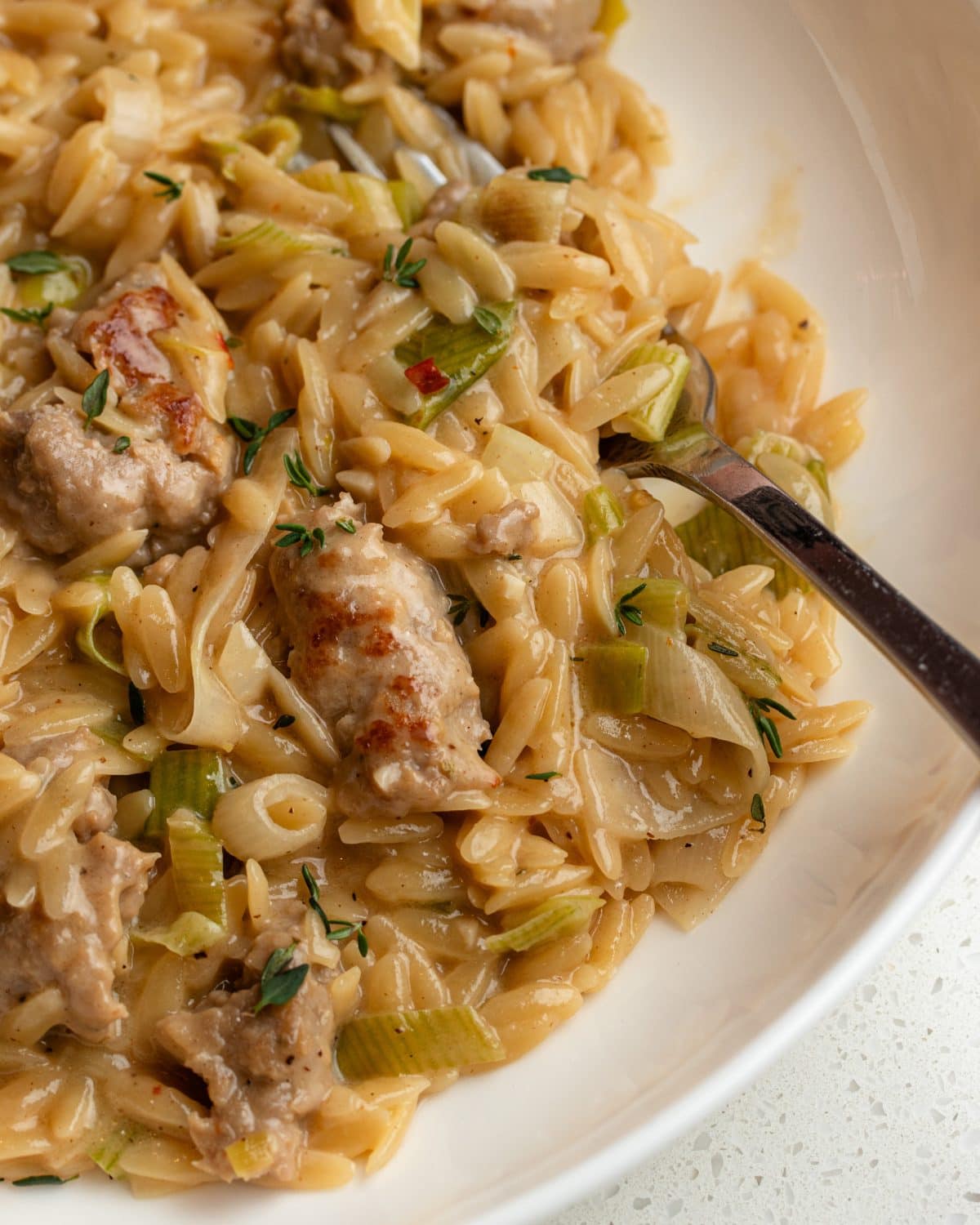 White plate with creamy orzo and sausage and leeks mixed through. A fork resting in.