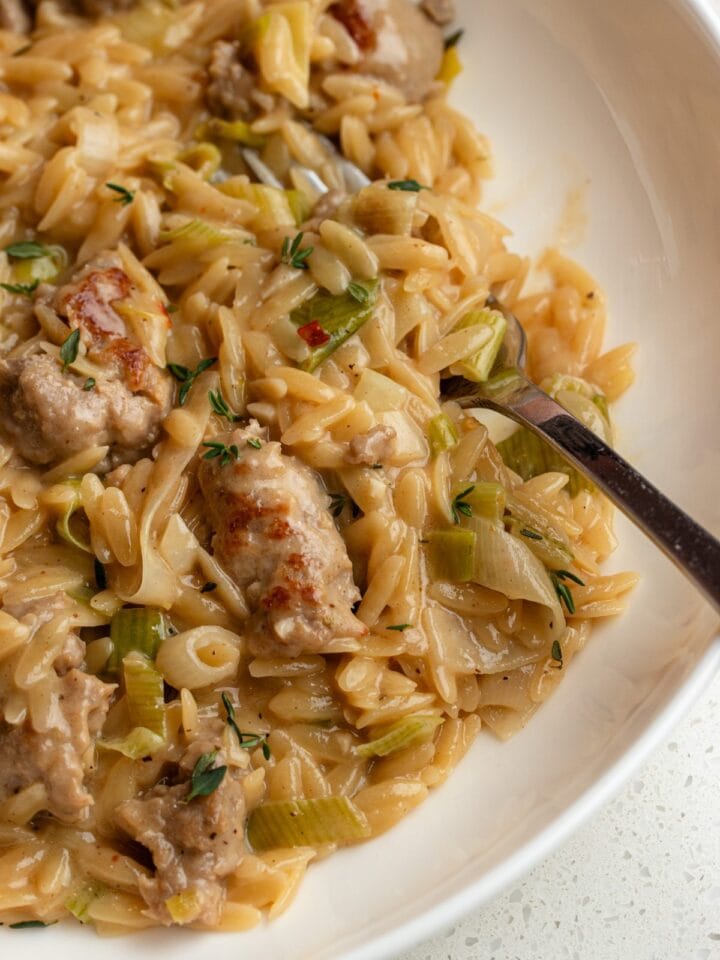 White plate with creamy orzo and sausage and leeks mixed through. A fork resting in.