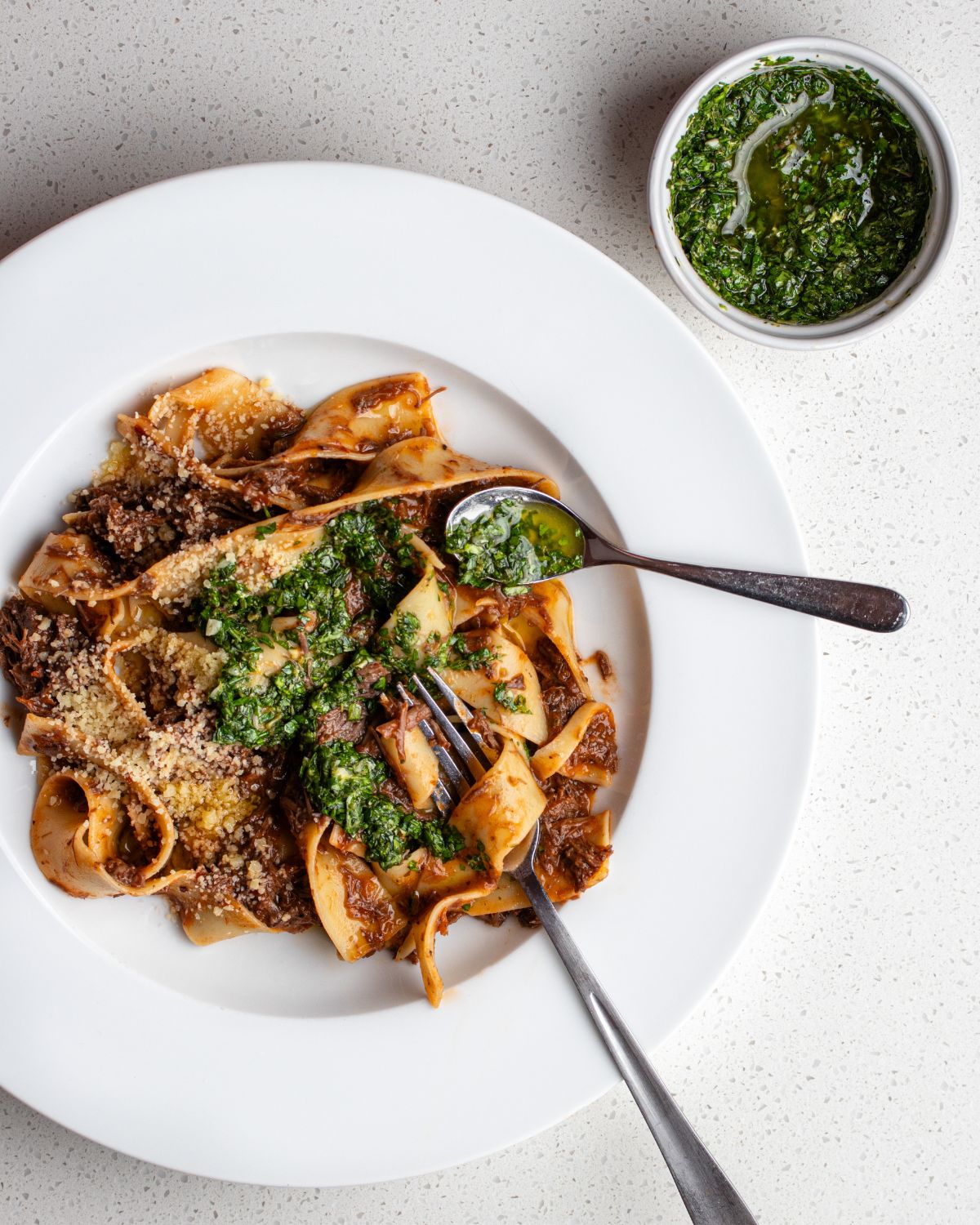 Gremolata drizzled into pappardelle pasta in a meat ragu. Ramekin with remaining gremolata above plate on bench.