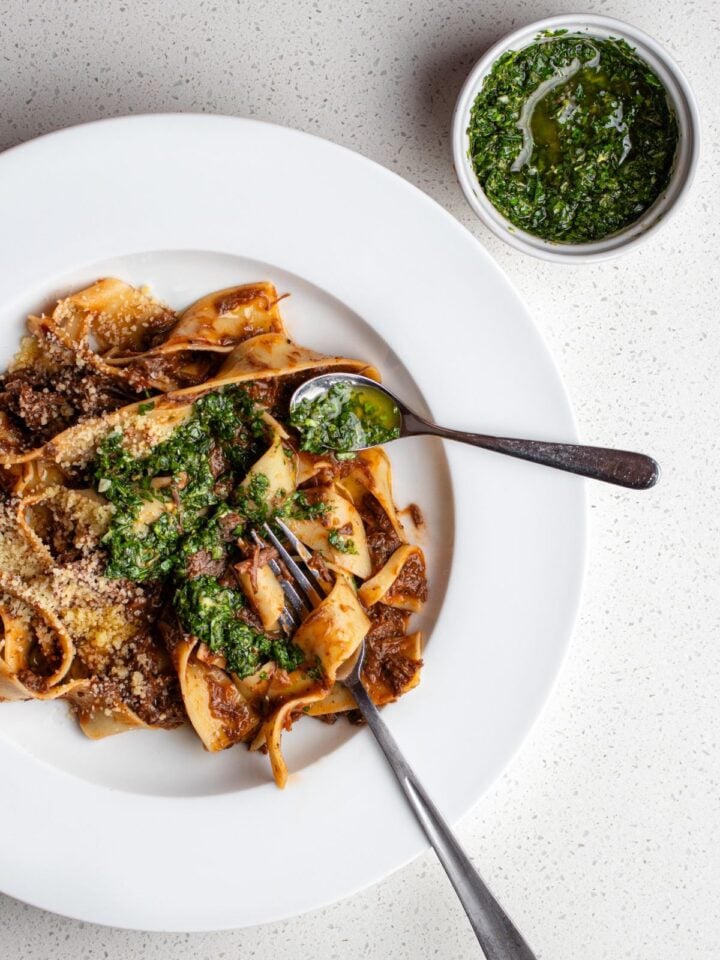 Gremolata drizzled into pappardelle pasta in a meat ragu. Ramekin with remaining gremolata above plate on bench.