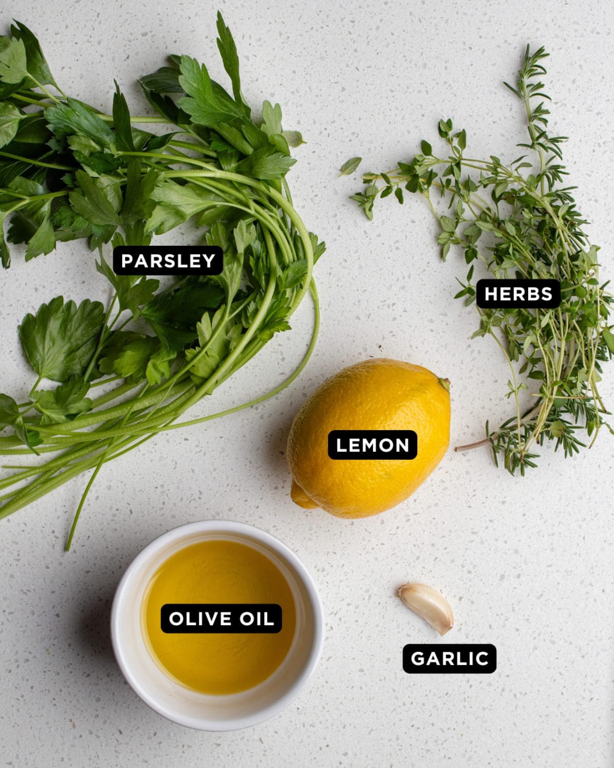 Gremolata ingredients on a bench top. Herbs, a lemon, a garlic clove and a ramekin of olive oil.