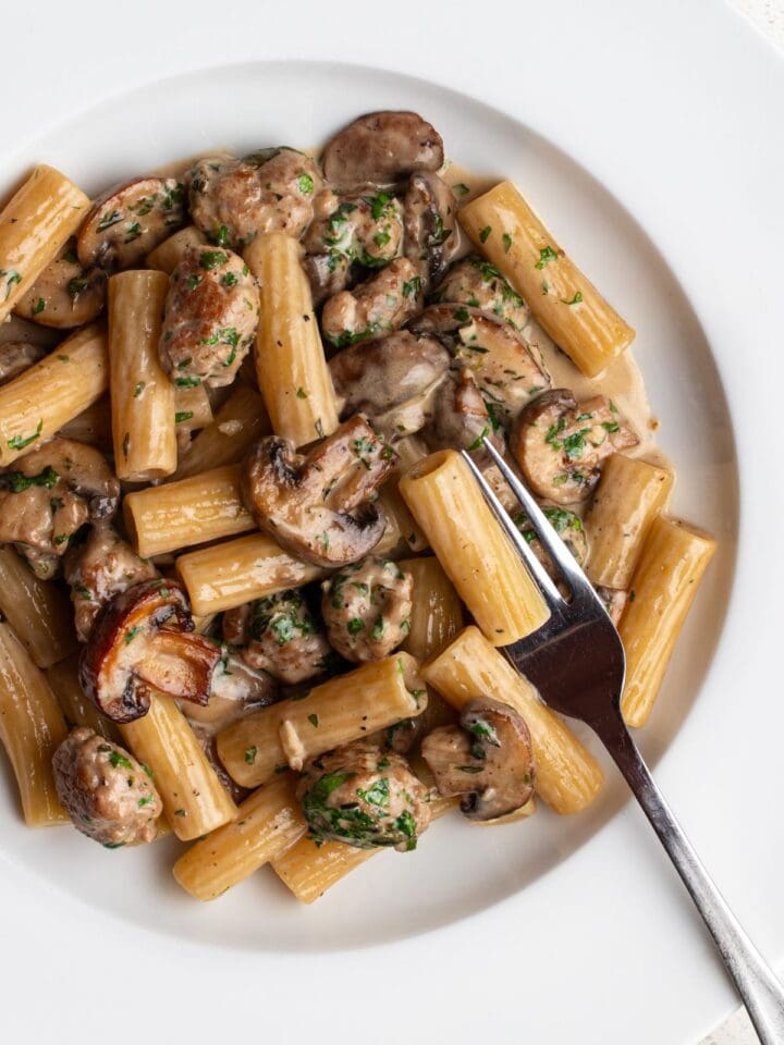 White plate with rigatoni in a sausage and mushroom sauce. Fork resting into pasta.