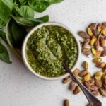 White bench with a ramekin of pistachip pesto. Scattered basil leaves and raw pistachios around it.