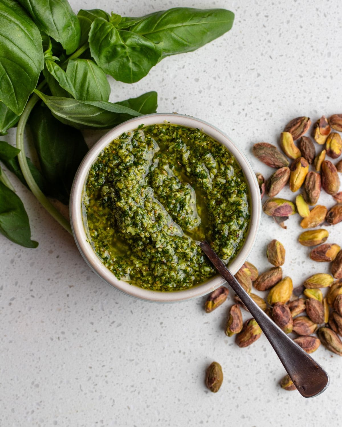 White bench with a ramekin of pistachip pesto. Scattered basil leaves and raw pistachios around it.