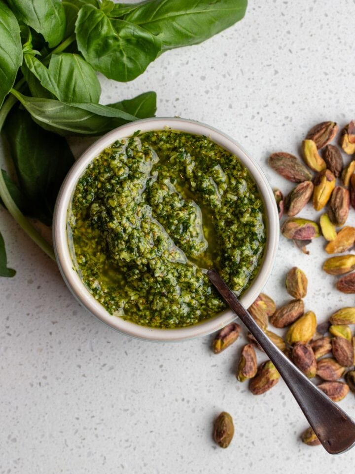 White bench with a ramekin of pistachip pesto. Scattered basil leaves and raw pistachios around it.