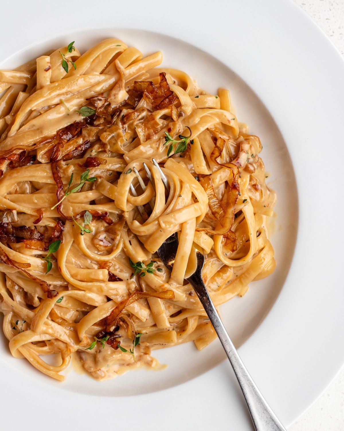 White plate with linguine in a creamy caramelized onion sauce. Fork resting onto plate.