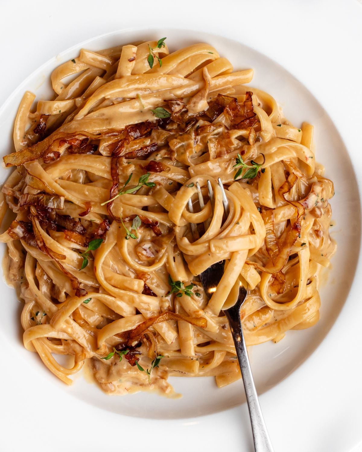 White plate with linguine in a creamy caramelized onion sauce. Fork resting onto plate.