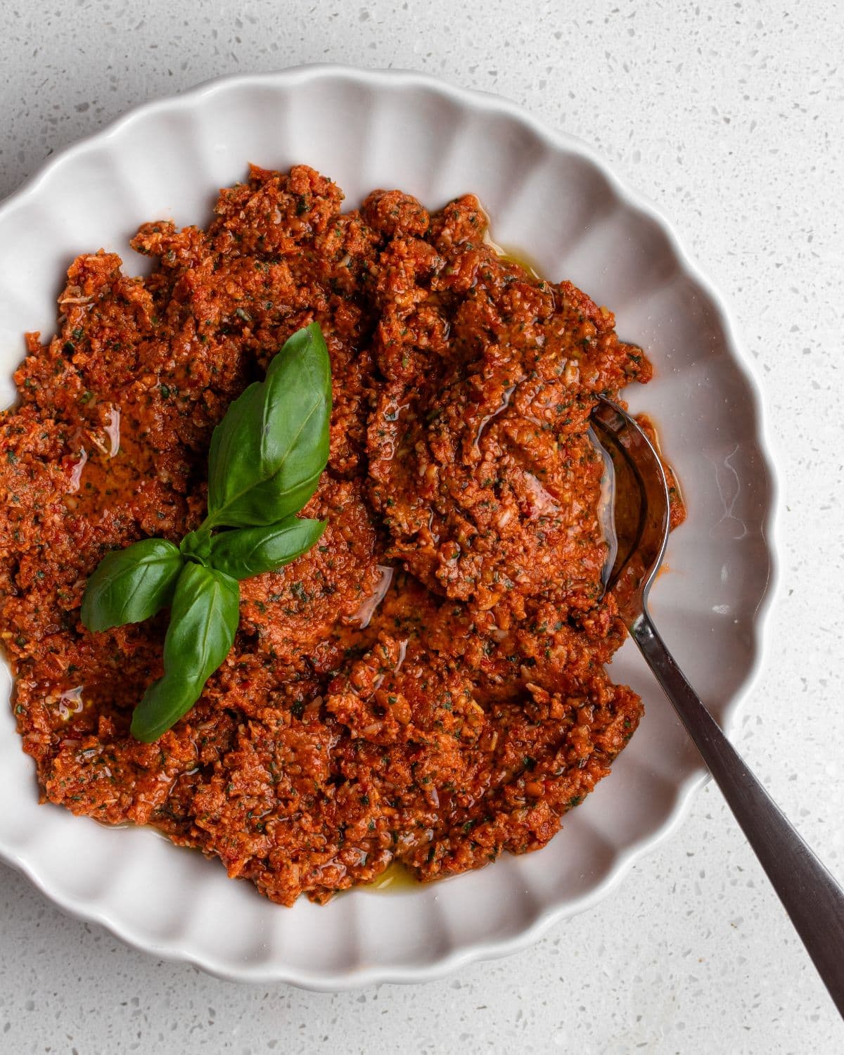 White bowl with sun-dried tomato pesto, a spoin resting in and basi leaves on top.