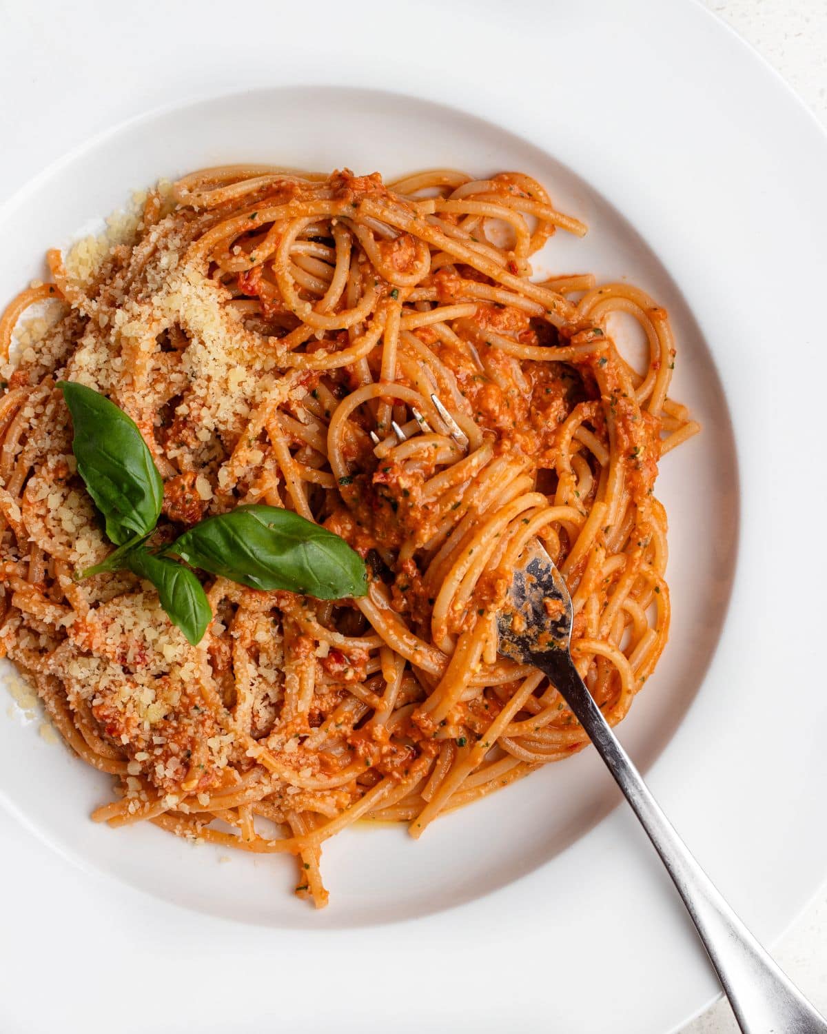 White plate with spaghetti in a sun-dried tomato pesto sauce. Basil on top and a fork resting in.
