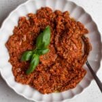 White bowl with sun-dried tomato pesto, a spoin resting in and basi leaves on top.