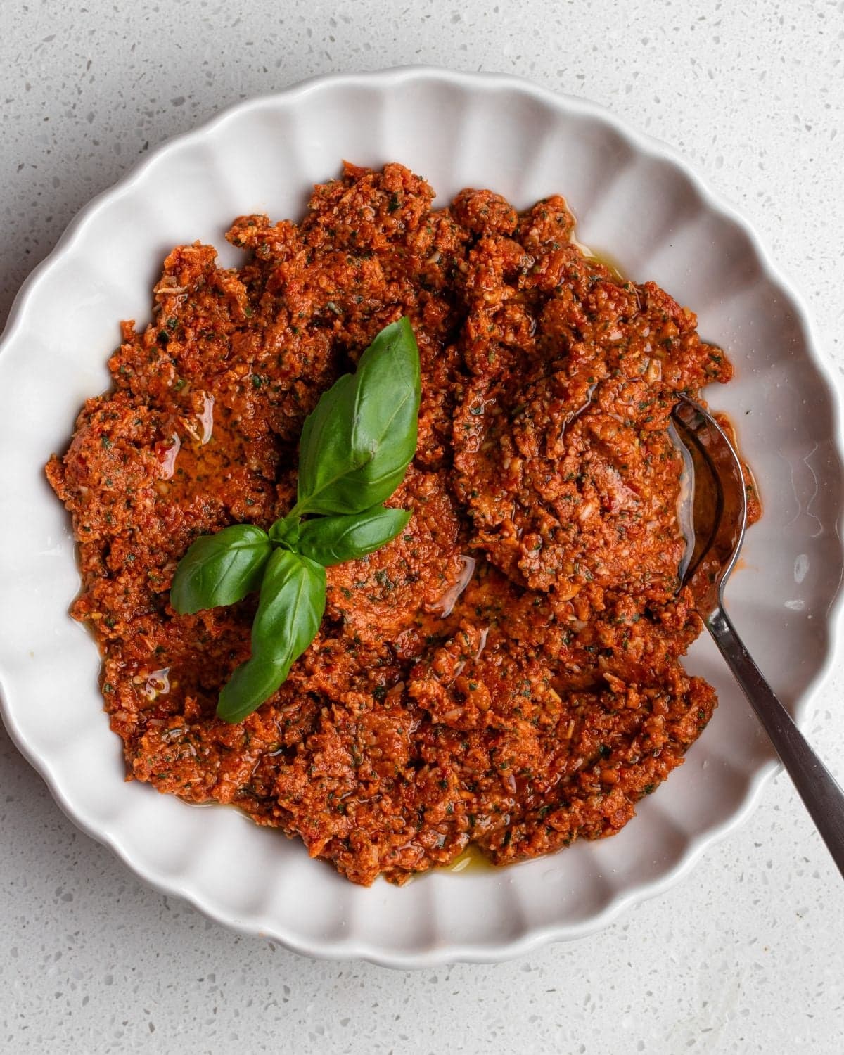 White bowl with sun-dried tomato pesto, a spoin resting in and basi leaves on top.