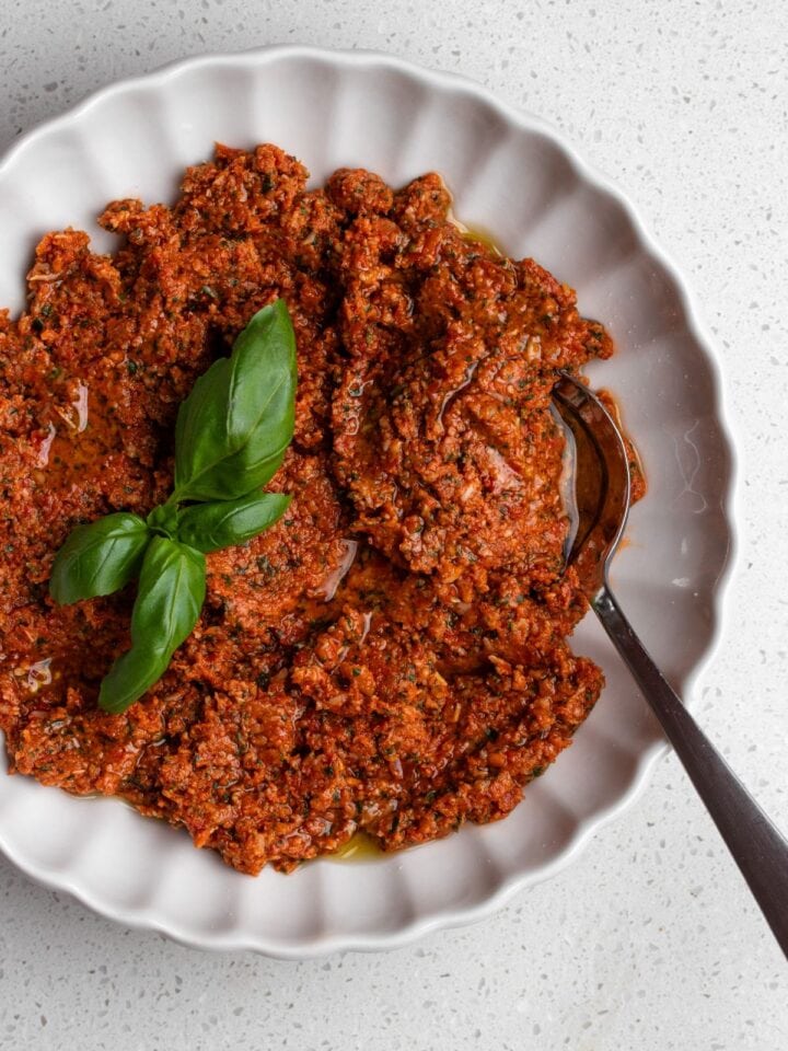 White bowl with sun-dried tomato pesto, a spoin resting in and basi leaves on top.