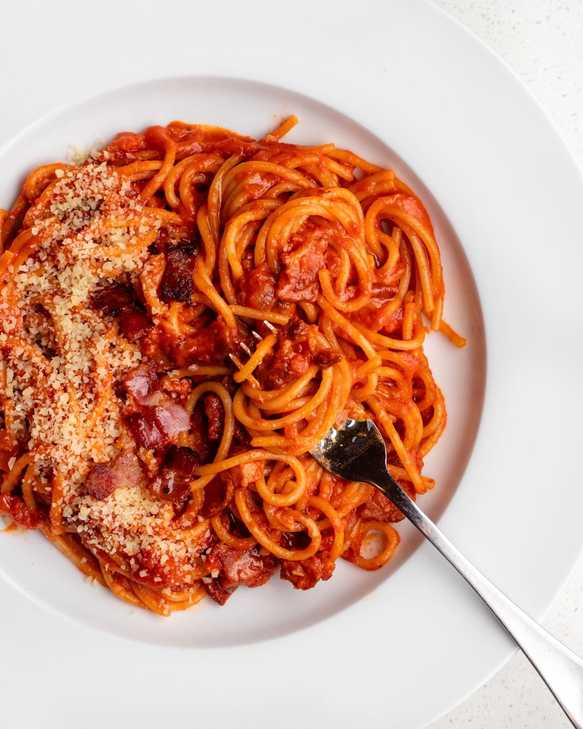 White plate with spaghetti in a tomato bacon pasta sauce with grated parmesan on top and a fork resting in.