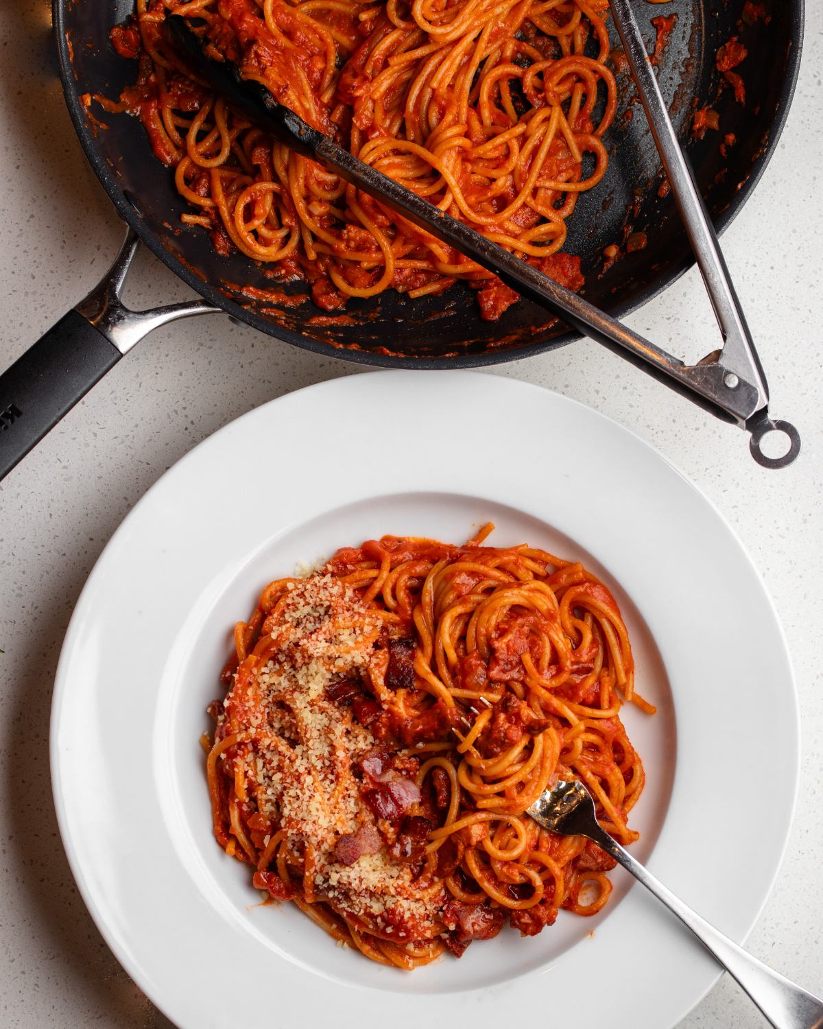White bench with a white plate with tomato bacon pasta. Skillet above with remaining pasta and tongs.