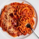 White plate with spaghetti in a tomato bacon sauce with grated parmesan on top and a fork resting in.
