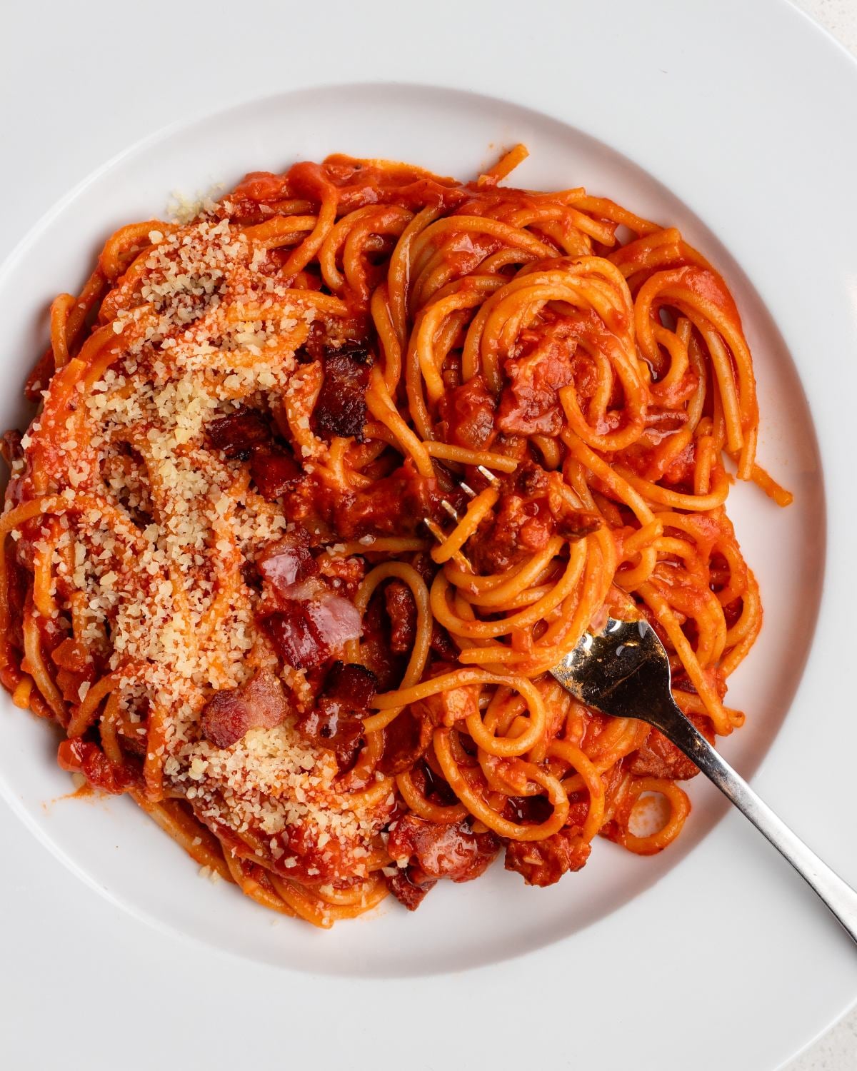 White plate with spaghetti in a tomato bacon sauce with grated parmesan on top and a fork resting in.