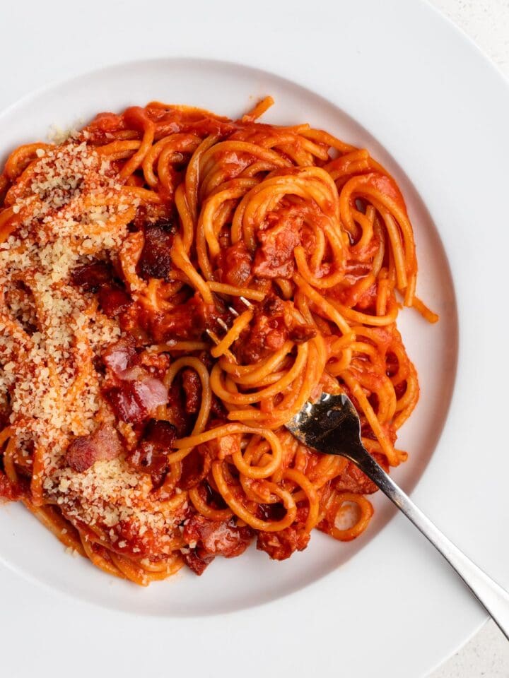 White plate with spaghetti in a tomato bacon sauce with grated parmesan on top and a fork resting in.