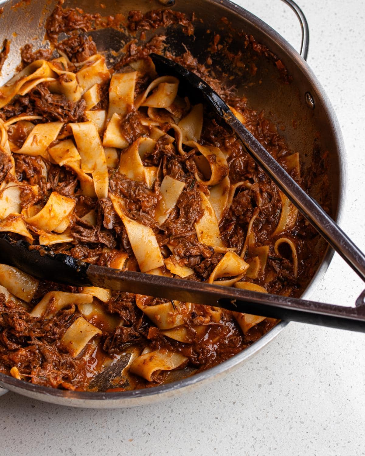 Skillet with pappardelle tossed in slow cooked beef ragu. Tongs resting in.