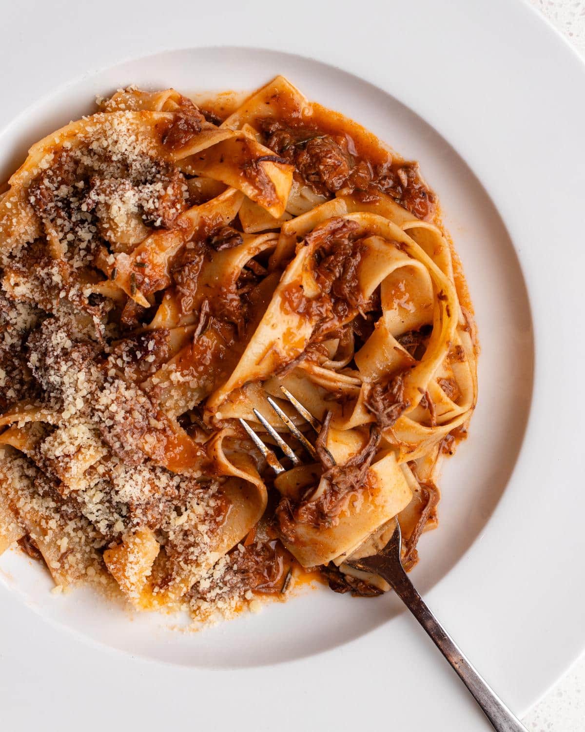 Beef ragu with pappardelle pasta on a white plate. Fork resting in and grated parmesan on top.