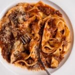 White plate with pappardelle pasta tossed in a slow cooked beef ragu. Fork resting in.