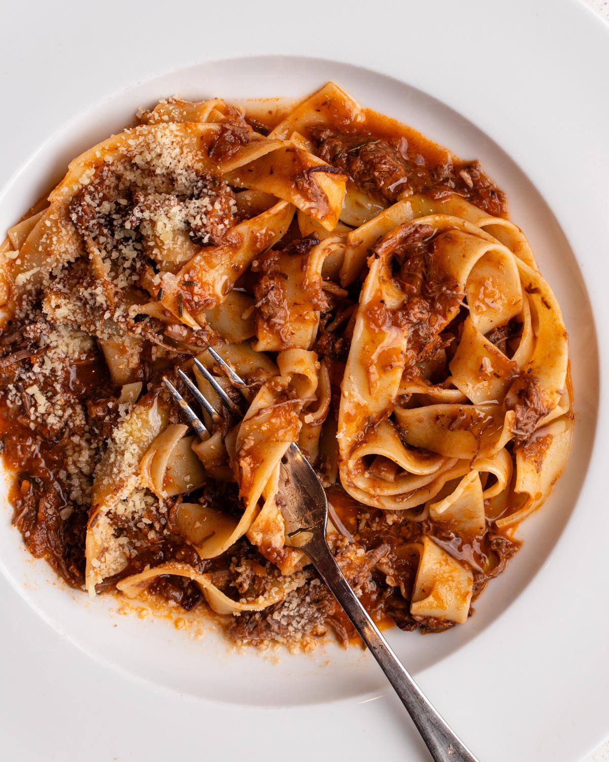 White plate with pappardelle pasta tossed in a slow cooked beef ragu. Fork resting in.