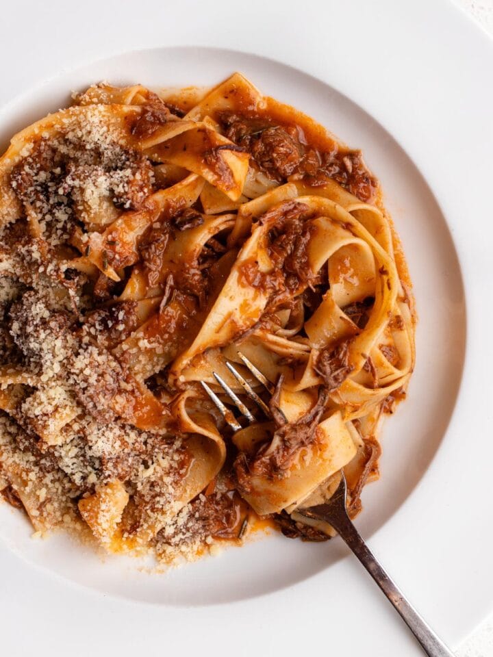 Beef ragu with pappardelle pasta on a white plate. Fork resting in and grated parmesan on top.