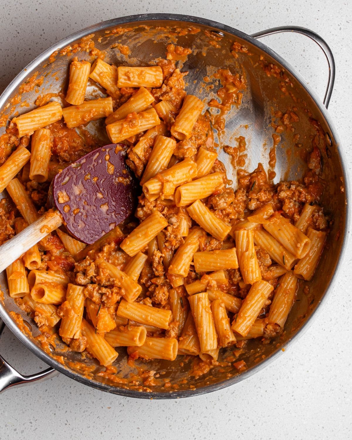 Stainless steel skillet with sausage ragu tossed theough rigatoni pasta. Spatula resting in.