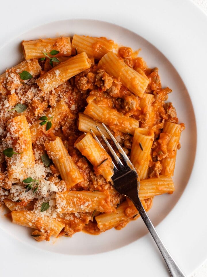 White plate with sausage ragu tossed through rigatoni. Fork resting in.
