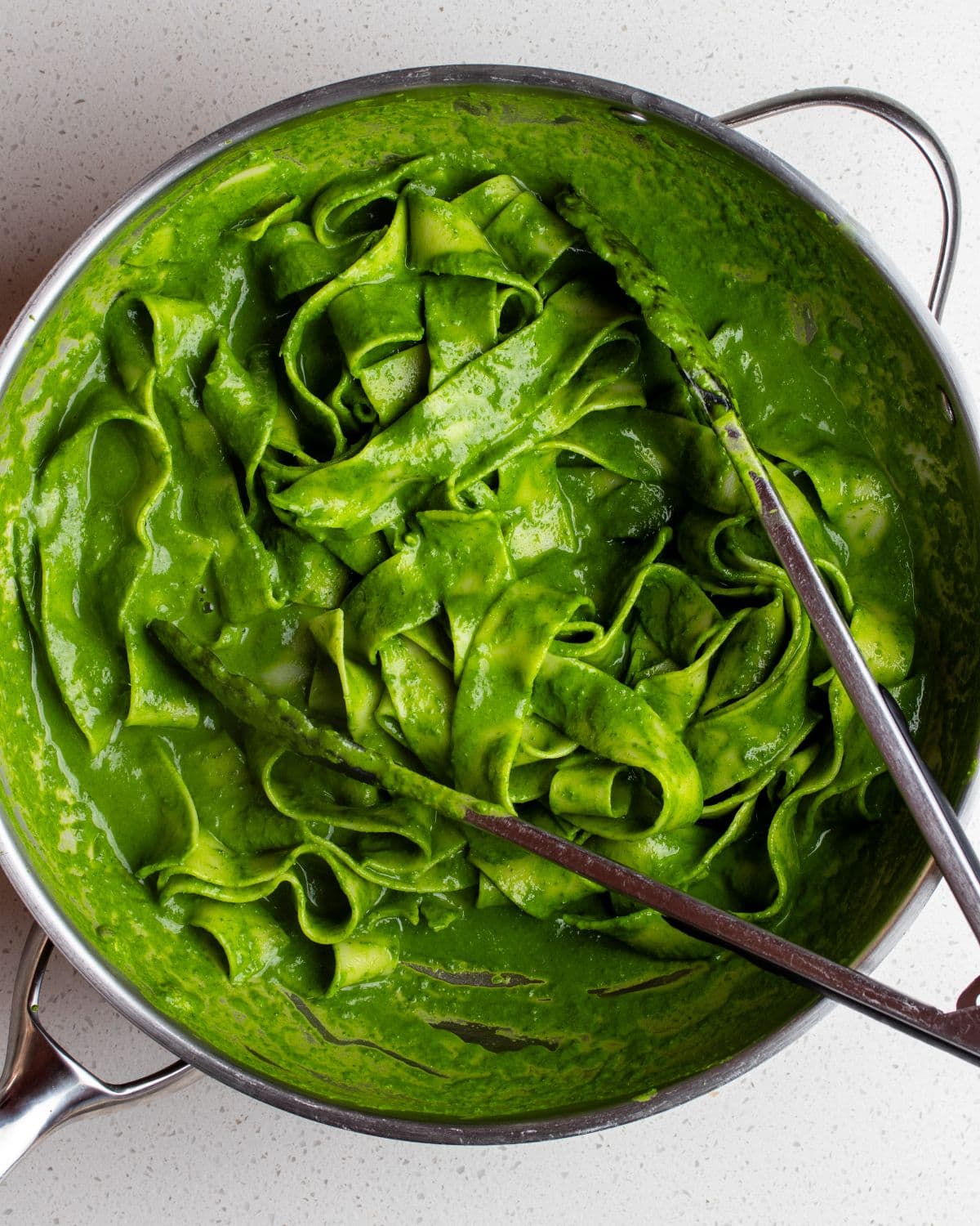 Stainless steel skillet with pappardell pasta in a vibrant green cavolo nero sauce. Tongs resting on top.