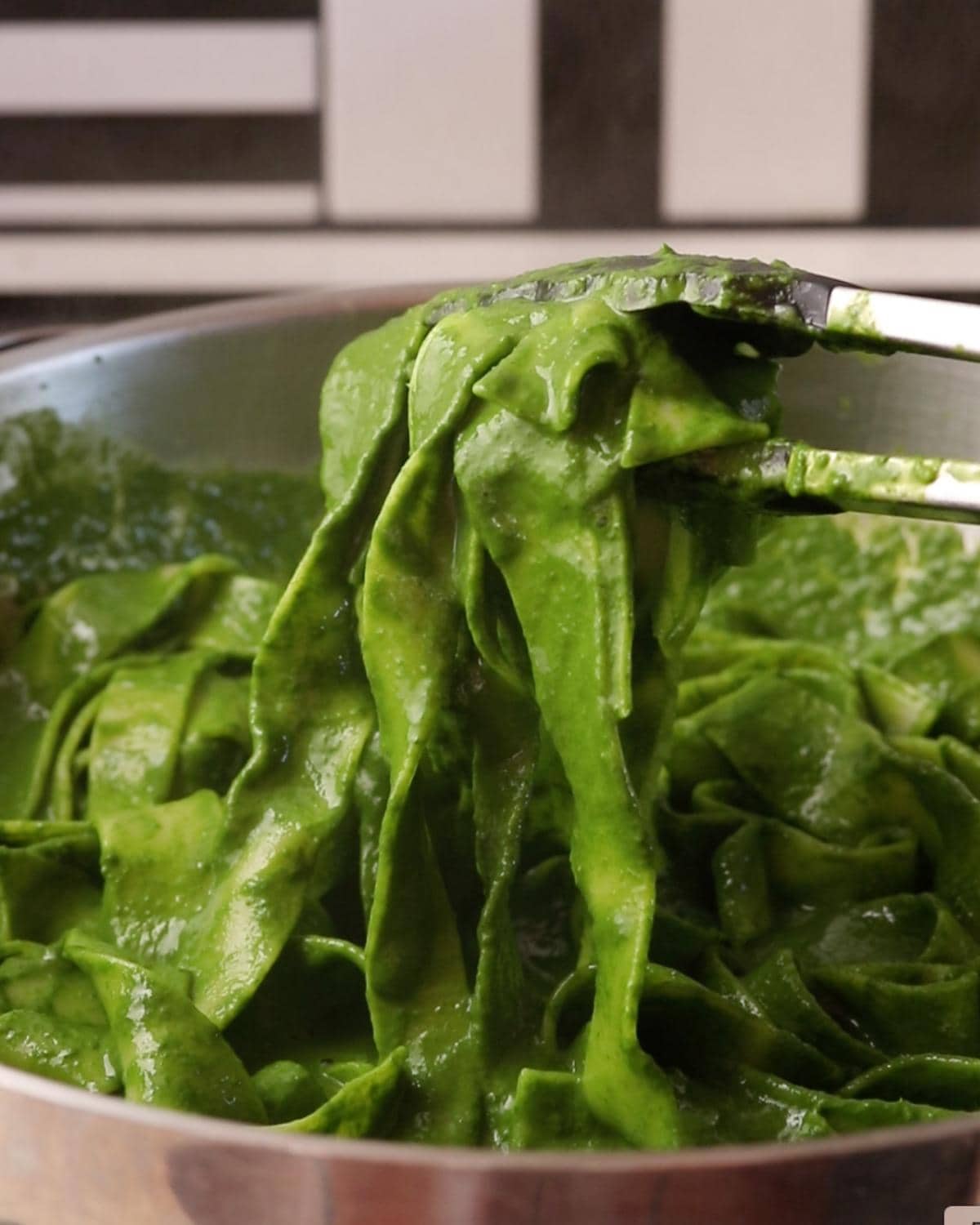 Pasta being tossed in a green cavolo nero sauce in a frying pan with tongs.